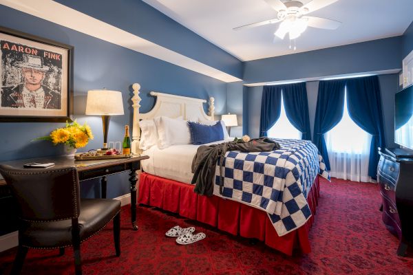 A cozy hotel room with a blue and white color scheme, a large bed, a desk, champagne, flowers, a painting, and a TV.