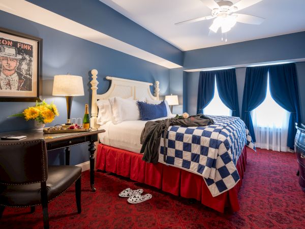 A cozy hotel room with a blue and white color scheme, a large bed, a desk, champagne, flowers, a painting, and a TV.