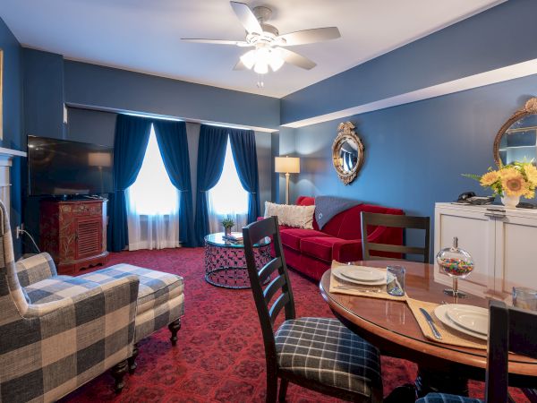 The image shows a cozy living room with blue walls, a red couch, patterned chairs, a dining table set, and a flat-screen TV, illuminated by natural light.