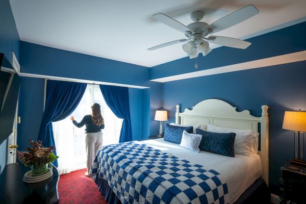 A person stands by a window in a blue bedroom with a checkered bedspread and ceiling fan, lamps, and a flower arrangement on a dresser.