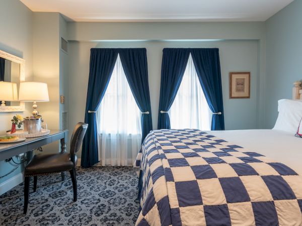 A cozy bedroom featuring a bed with a checkered quilt, a vanity with a lamp and mirror, two windows with blue curtains, and a patterned carpet.