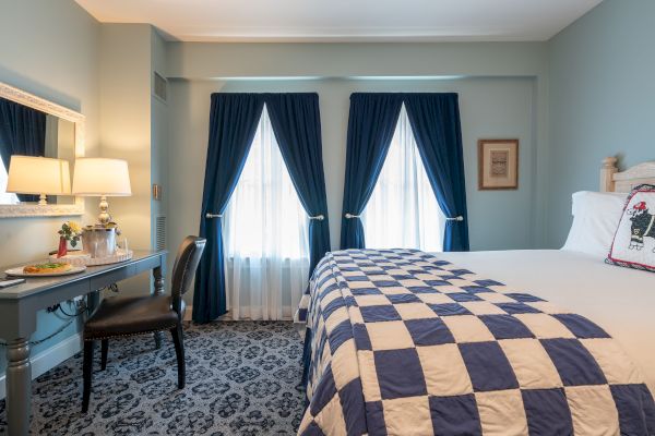 A cozy bedroom featuring a bed with a checkered quilt, a vanity with a lamp and mirror, two windows with blue curtains, and a patterned carpet.