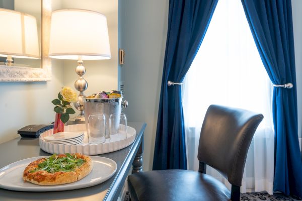 A room with a desk setup including a lamp, flowers, drinks, and a plate of food near a chair in front of a window with blue curtains.