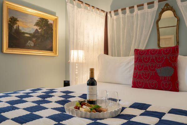 A cozy room with blue walls and white furniture, featuring a framed picture, a desk with a lamp, flowers, champagne, and a bed with a checkered blanket.