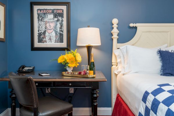 A cozy room with blue walls and white furniture, featuring a framed picture, a desk with a lamp, flowers, champagne, and a bed with a checkered blanket.