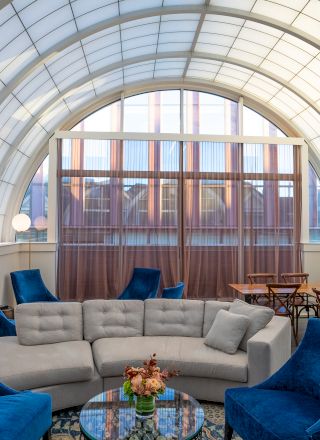 A modern lounge with blue chairs, a gray sofa, a glass coffee table, and globes on shelves, under an arched, light-filled ceiling.