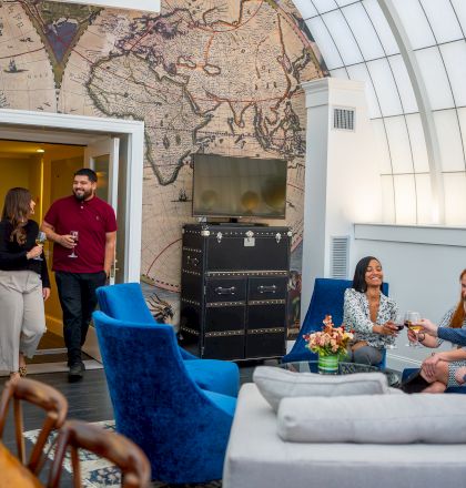 People socializing in a room with a vintage map mural, blue chairs, a TV, and a globe.