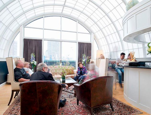 People are seated in a bright, arched-window room with a bar and flower arrangements in the background.