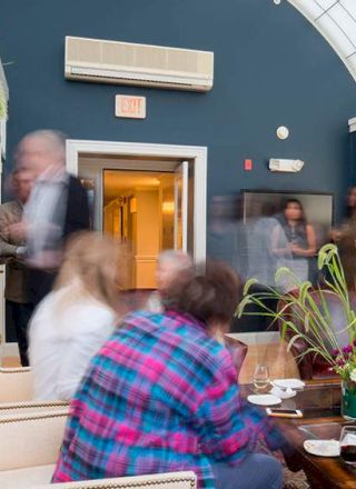 A group of people are gathered in a lounge with blue walls, a large window, and a plant on a coffee table.