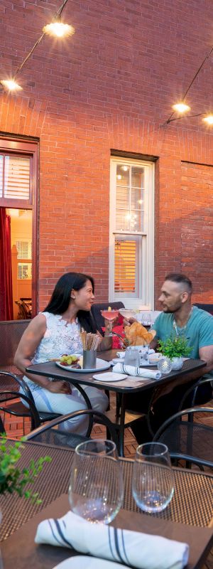 A couple is dining outdoors at a restaurant with brick walls, under string lights and large umbrellas.