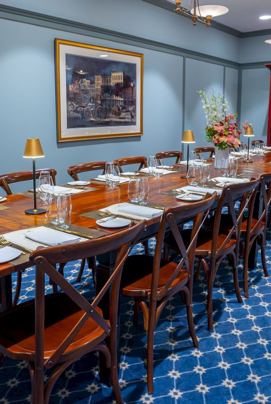 A well-set dining room with a long wooden table, chairs, elegant tableware, modern lighting, a blue patterned carpet, and a display cabinet.