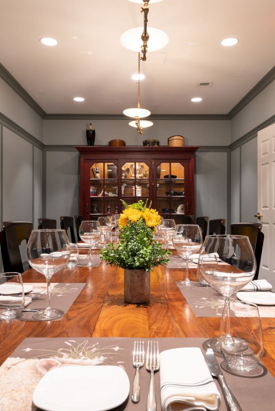 A dining room features a long wooden table set for a formal meal, complete with plates, cutlery, wine glasses, and a centerpiece of yellow flowers.