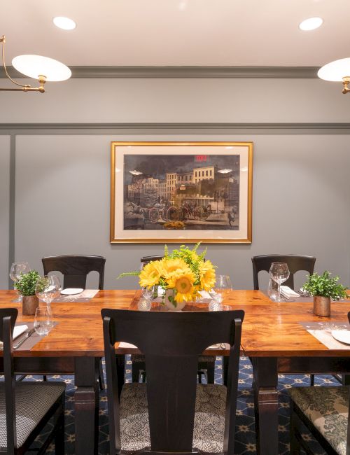 The image shows a dining room with a long wooden table, set with plates and glasses, decorated with plants and a centerpiece of yellow flowers.