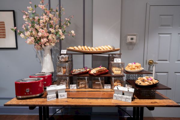 A table with an assortment of pastries, toasters, and packed food items, with a vase of flowers in the background and a door on the right.