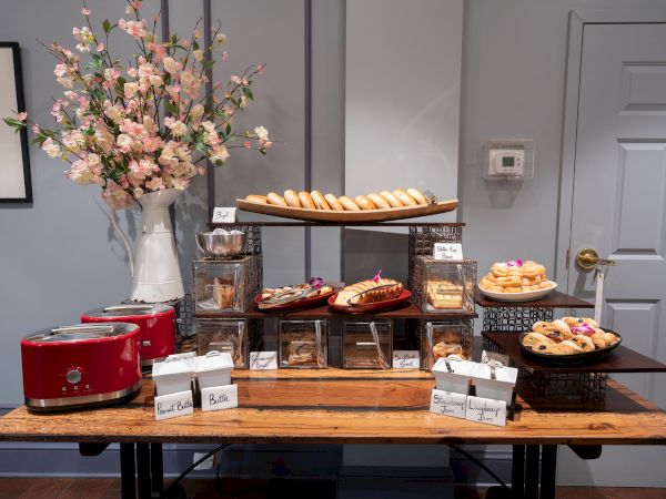 A table with an assortment of pastries, toasters, and packed food items, with a vase of flowers in the background and a door on the right.