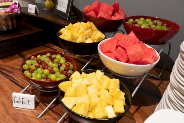 Bowls of fresh fruit on a table, including grapes, pineapple, and watermelon, with a sign labeled 