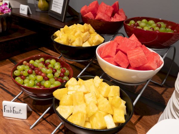 Bowls of fresh fruit on a table, including grapes, pineapple, and watermelon, with a sign labeled "Fresh Fruit." Plates are also nearby.
