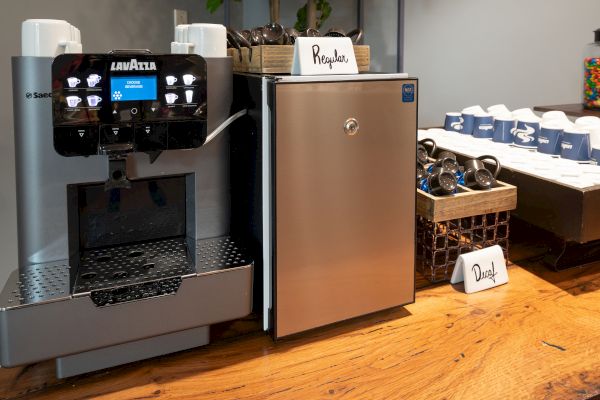 The image shows a coffee station featuring a Lavazza coffee machine, a small refrigerator labeled 'Regular,' and cups and lids arranged neatly on a counter.