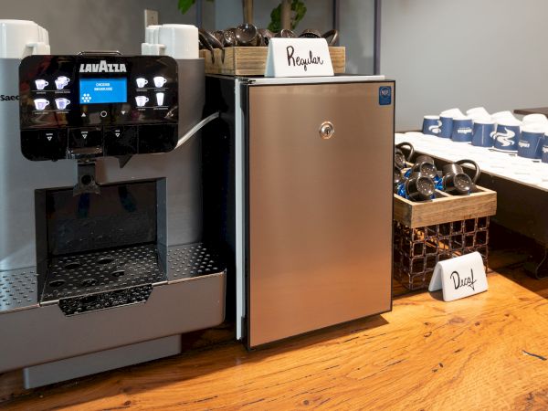 The image shows a coffee station featuring a Lavazza coffee machine, a small refrigerator labeled 'Regular,' and cups and lids arranged neatly on a counter.
