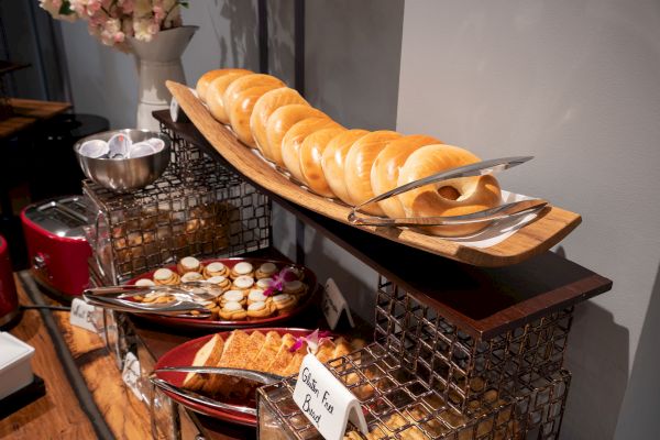 The image shows a buffet display with bagels on a wooden platter, tongs, a bowl of toppings, and other assorted baked goods on separate plates.