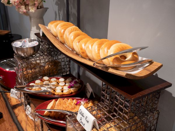 The image shows a buffet display with bagels on a wooden platter, tongs, a bowl of toppings, and other assorted baked goods on separate plates.