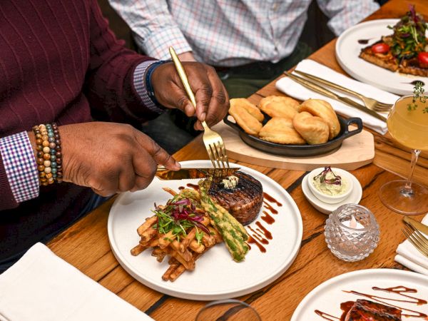 A person is about to eat a meal of steak, fries, and asparagus, with rolls, butter, and a drink visible on the table.