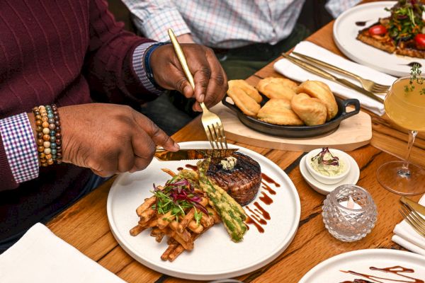 A person is about to eat a meal of steak, fries, and asparagus, with rolls, butter, and a drink visible on the table.