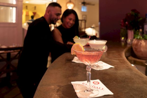 Two people at a bar with cocktails placed on napkins. The setting has warm lighting and a cozy atmosphere.