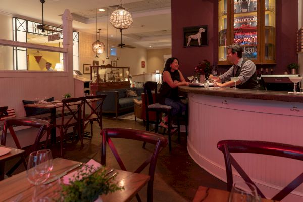 A cozy restaurant bar with two people conversing, wooden tables and chairs, and warm lighting creating an inviting atmosphere.