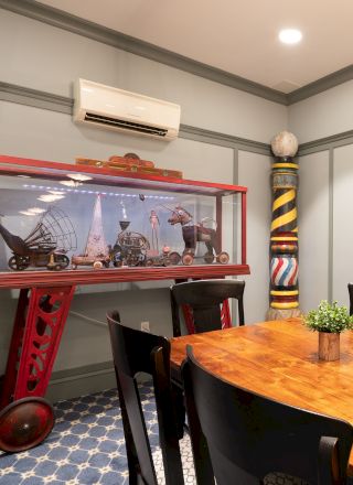 A wooden table with black chairs, a display case with model ships, and a colorful post are in a room with gray walls and a patterned floor.