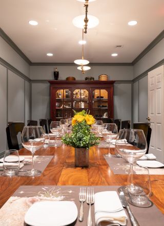 A dining room with a long wooden table set for dinner, featuring a centerpiece with yellow flowers, overhead lighting, and wall decor.