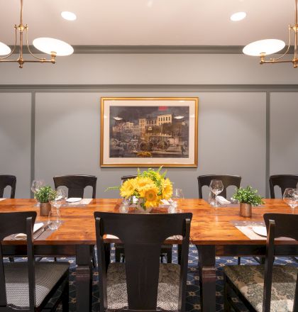 A dining room setup with a wooden table, chairs, flower centerpiece, framed artwork on the wall, and ceiling lights, all elegantly arranged.