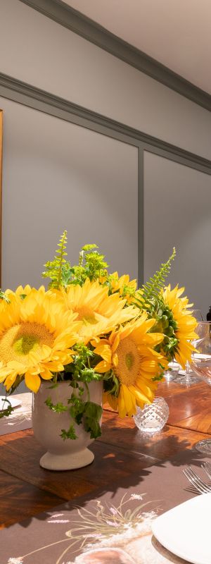 A dining room with a wooden table set for a meal, adorned with yellow flowers in the center, and a glass cabinet filled with items in the background.