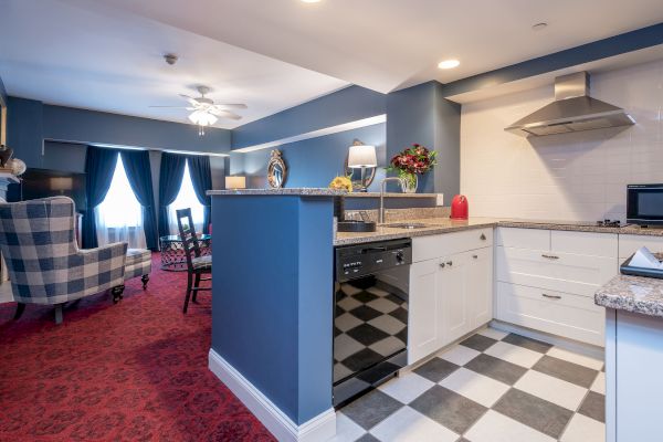 The image shows a modern kitchen with white cabinets, checkered flooring, and an adjoining living area with red carpet, blue walls, and a chair.