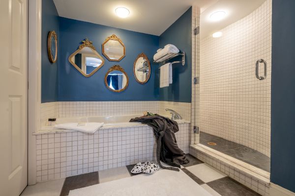 A bathroom with blue walls, vintage mirrors, a bathtub, shower, towel rack, robe, slippers, and tiled walls and floor.