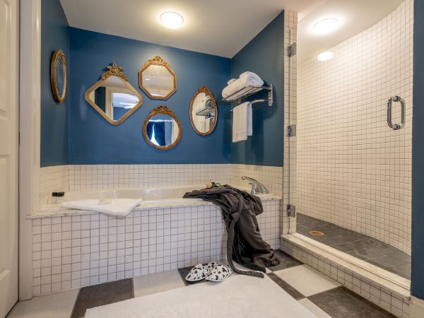 A bathroom with blue walls, vintage mirrors, a bathtub, shower, towel rack, robe, slippers, and tiled walls and floor.