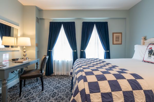 The image shows a neatly arranged bedroom with a bed covered in a blue and white checkered quilt, a desk with a lamp and chair, and two windows.