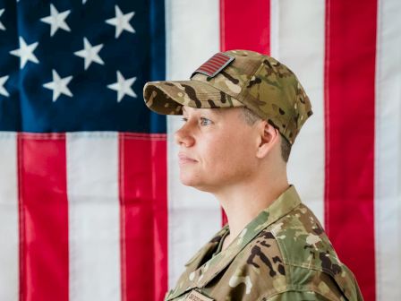 A person in military uniform stands in front of an American flag, looking to the side, with a serious expression on their face.