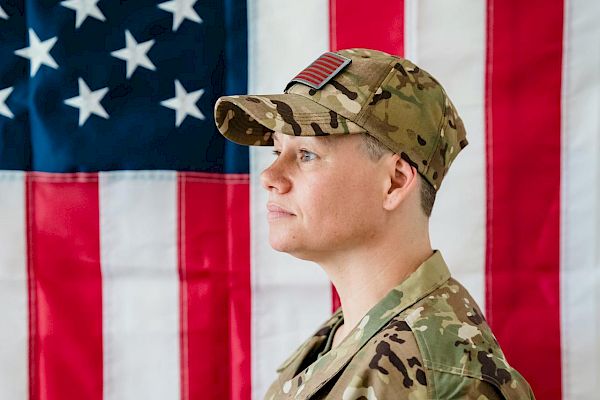 A person in military uniform stands in front of an American flag, looking to the side, with a serious expression on their face.