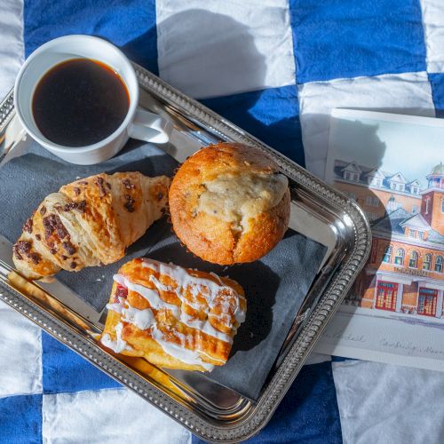 A tray with coffee, a croissant, muffin, pastry, and an illustration on a blue and white checkered background.