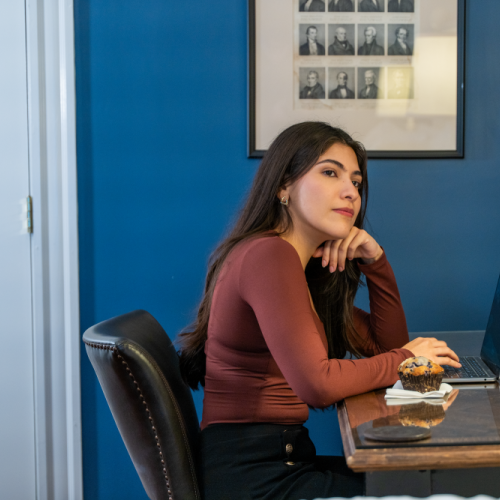 A woman sits at a desk in a blue-walled office, resting her head on her hand, with a laptop and a pastry in front of her.