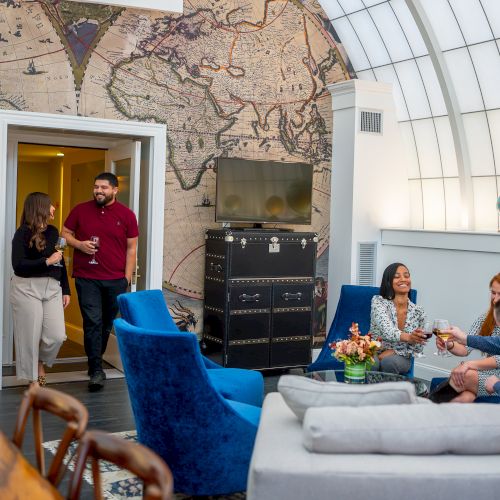 A group of people socializing in a stylish room with a world map wall, blue chairs, and a decorative globe display.