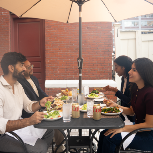 A group of four friends enjoys a sunny outdoor meal at a patio table with salads, drinks, and a shared conversation under a large umbrella.