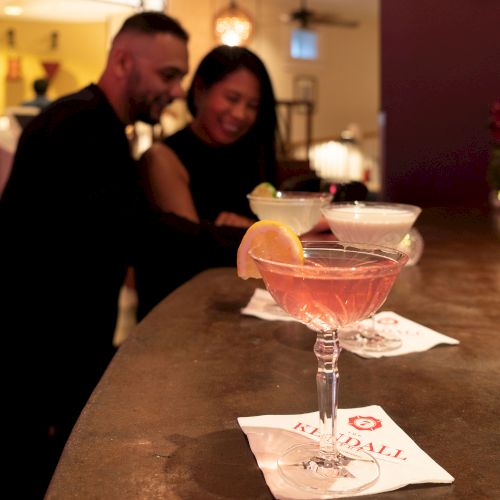 A couple is standing at a bar with three cocktails on the counter, each garnished with fruit and placed on napkins with the logo "George."