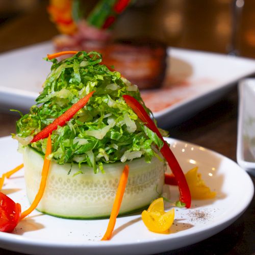 A creatively presented salad features shredded lettuce, cucumber, bell peppers, carrots, and sliced cherry tomatoes on a white plate.