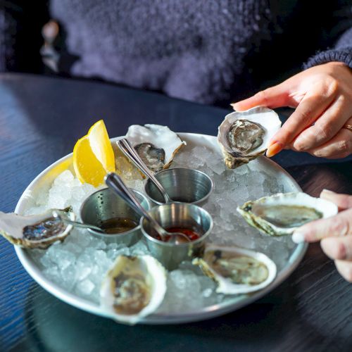 A plate of oysters on ice with lemon slices and sauces, accompanied by a glass of white wine, and hands reaching for the oysters.