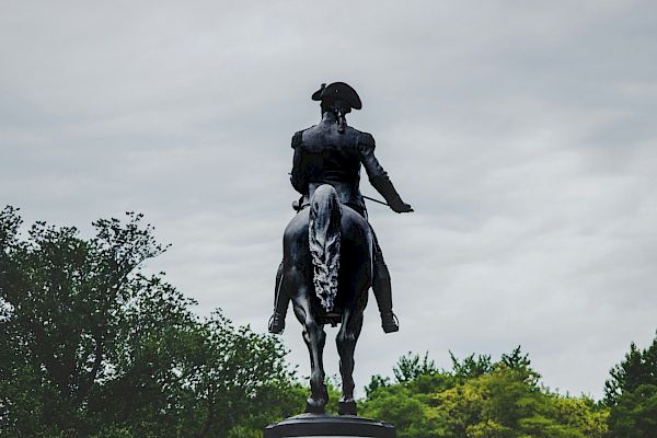 The image shows a statue of a person on horseback atop a pedestal in a park setting, with trees and a cloudy sky in the background.