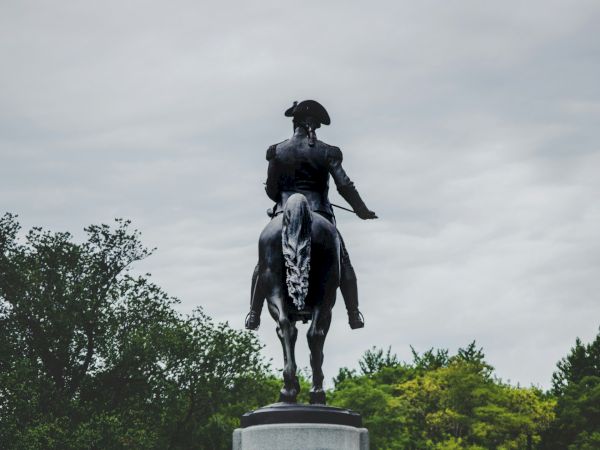 The image shows a statue of a person on horseback atop a pedestal in a park setting, with trees and a cloudy sky in the background.
