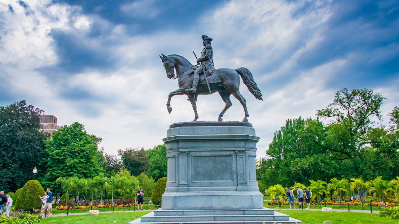 The image shows an equestrian statue in a park surrounded by greenery, flowers, and people walking around in the background.