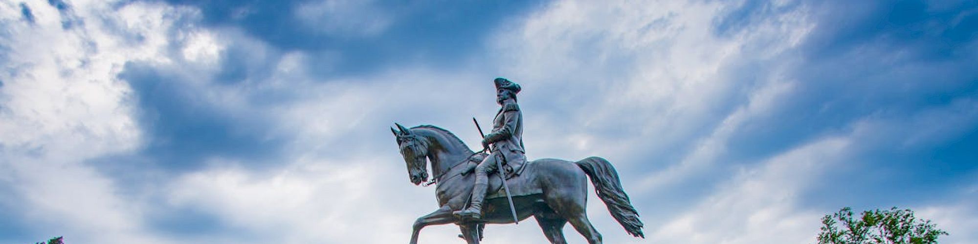 The image shows an equestrian statue in a park surrounded by greenery, flowers, and people walking around in the background.